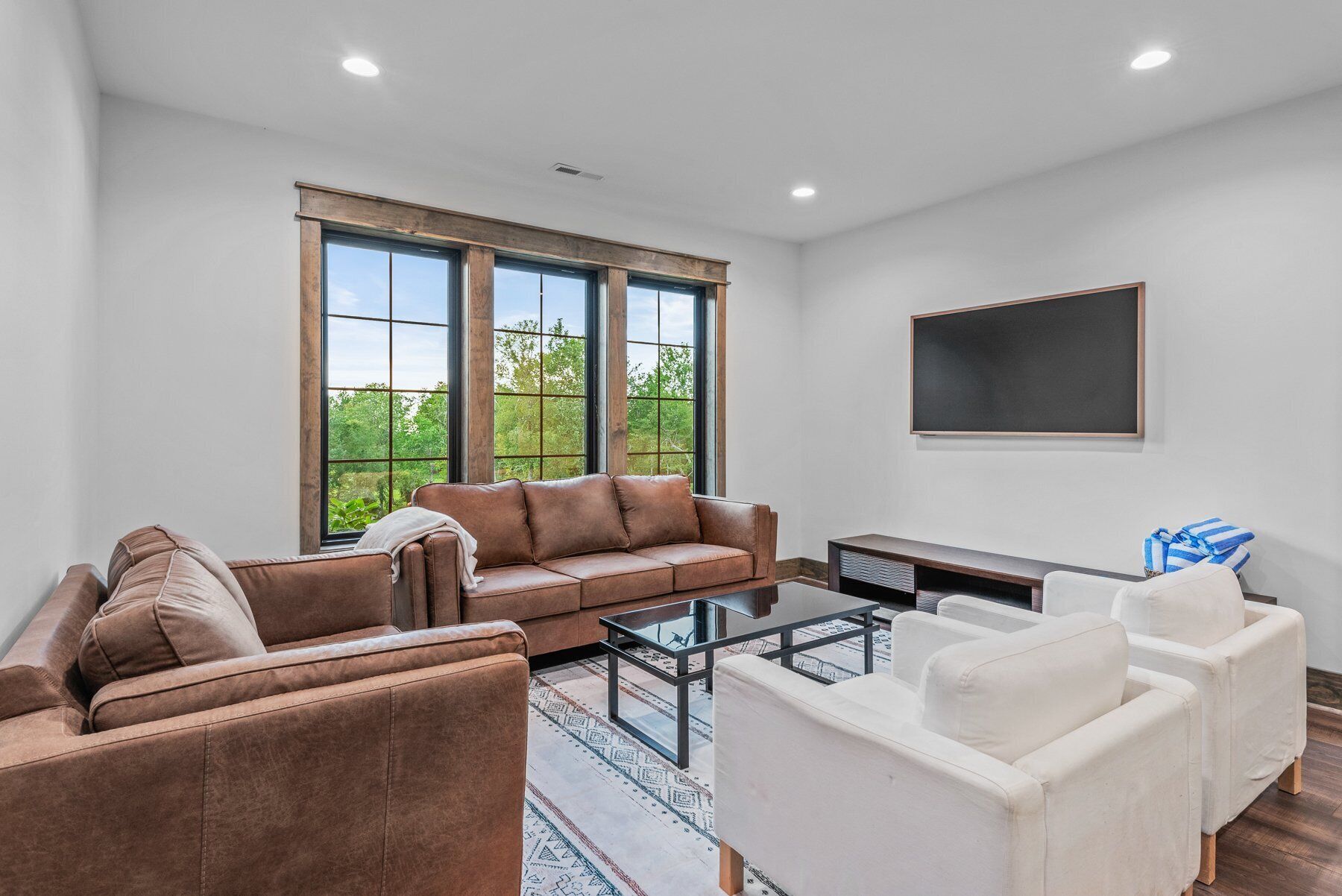 A spacious living room with a brown leather sofa, a white armchair, and a glass coffee table. Large windows offer a view of greenery outside.