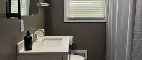 Een badkamer met een wit wastafel, toilet, raam met zonwering, douchegordijn en spiegel.