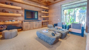 A cozy log cabin interior with a large TV, wooden shelves, comfortable seating, and a view of greenery outside.