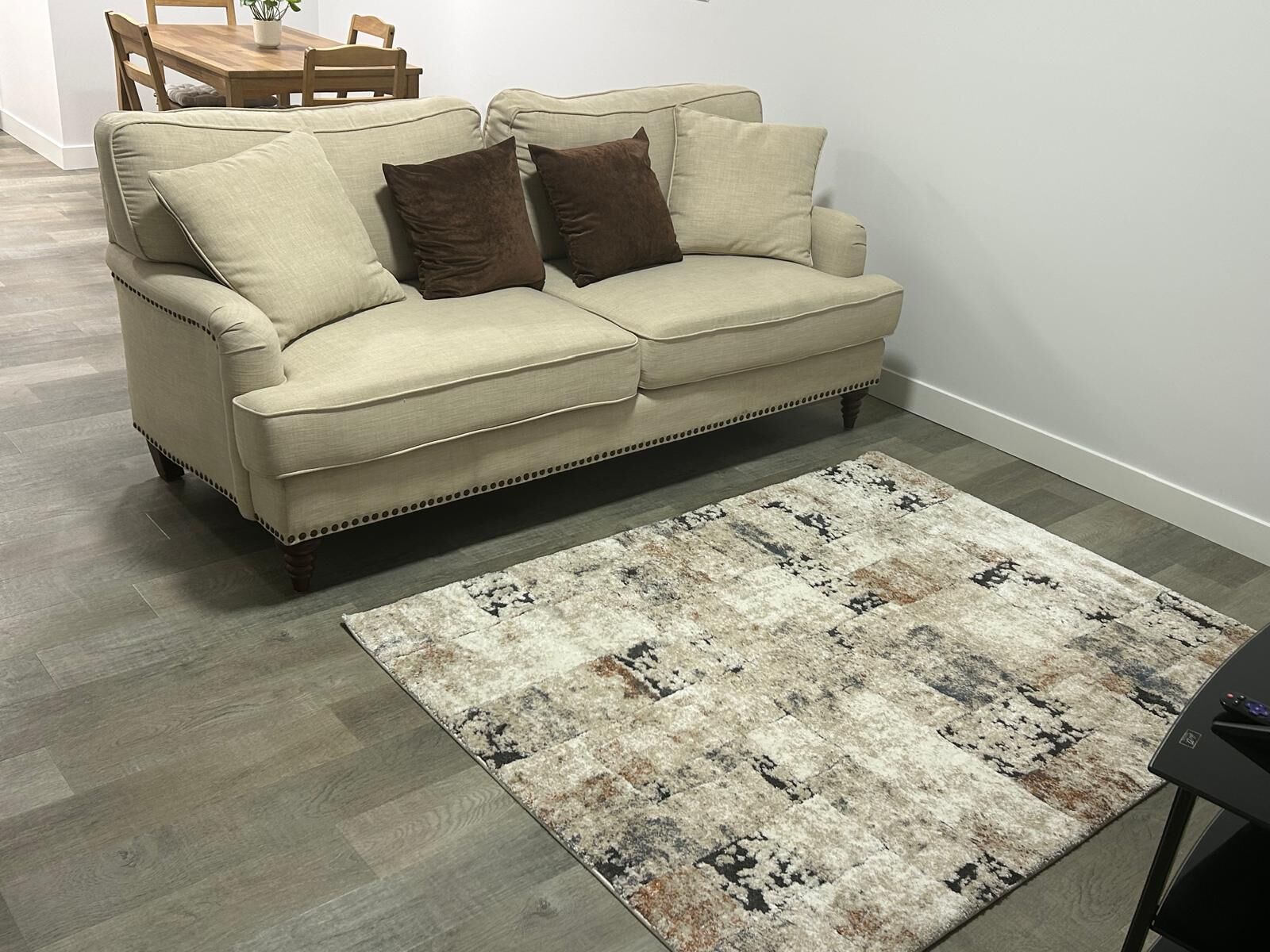 Ein beiges Sofa mit braunen Kissen, ein gemustertes Teppich und ein Holztisch mit Stühlen im Hintergrund.