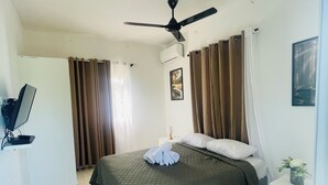 Ein Hotelzimmer mit einem Bett, einem Deckenventilator, einer Klimaanlage, einem Nachttisch mit Lampe und einer kleinen Pflanze.