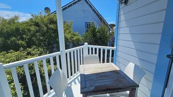 A wooden table and white chair on a porch with a white railing, overlooking a blue house and greenery.