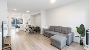Un salon moderne avec un canapé d’angle gris, une table à manger avec des chaises et une grande fenêtre qui laisse entrer la lumière naturelle.