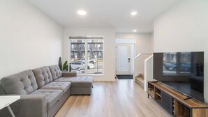 Un salon moderne avec un canapé d’angle gris, un téléviseur à écran plat posé sur un meuble en bois et une grande fenêtre donnant sur des voitures garées et des immeubles.
