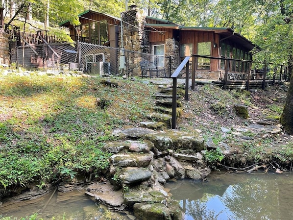 A rustic cabin with a stone wall and a wooden deck, surrounded by a natural landscape with a small stream. - Oak Shores Oasis on Lake Ouachita! (Mount Ida)