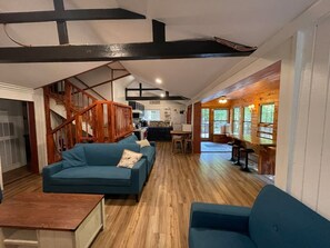 A spacious living area with a blue sofa, wooden flooring, a staircase, and a kitchen in the background. - Oak Shores Oasis on Lake Ouachita! (Mount Ida)