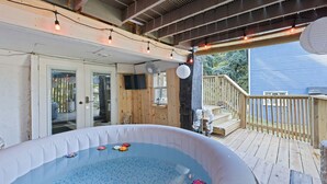 An outdoor hot tub area with a wooden deck, a covered seating area, and a view of a house in the background.