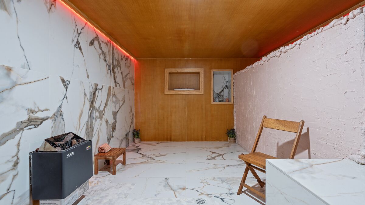 A sauna room with wooden walls, a wooden ceiling, and a marble floor. There is a wooden chair, a small wooden stool, and a heater with a box labeled "YETEX".