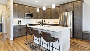 A modern kitchen with wooden cabinets, stainless steel appliances, a white island with bar stools, and pendant lighting.