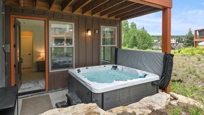 A hot tub outside a wooden building with a glass door, a stone ledge, and a view of a forested area.