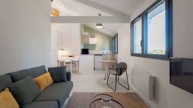 A modern living room with a grey sofa, a coffee table, and a dining area with a table and chairs. A kitchen is visible in the background with a window and a view of greenery outside. - Lovely country T2 near town center! (Aix-en-Provence)