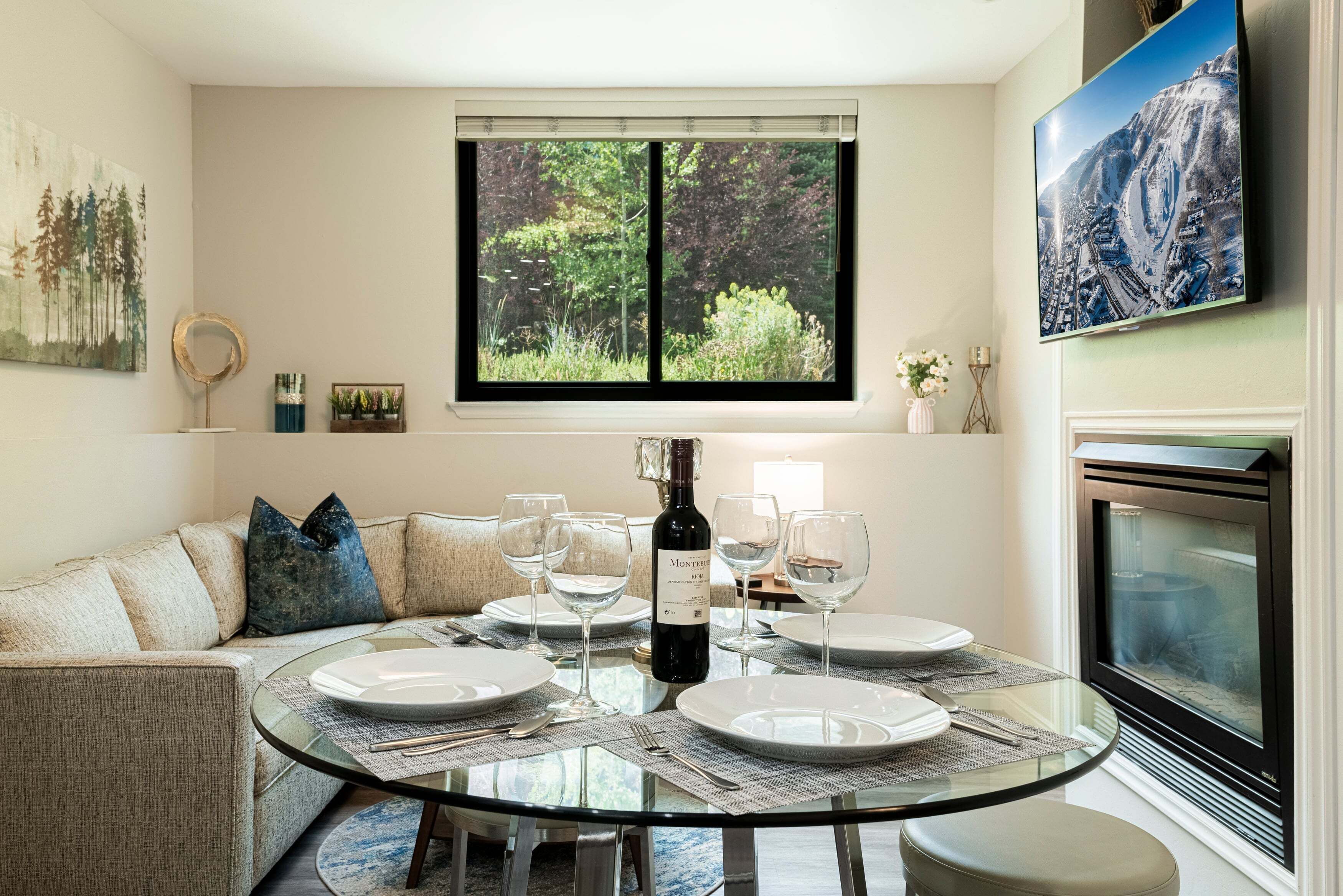 A dining area with a round glass table set for four, a bottle of wine, and a television mounted on the wall.
