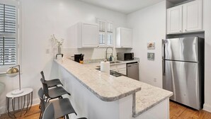 A modern kitchen with white cabinets, a stainless steel refrigerator, a marble countertop, and a small island with bar stools. - Retreat on Spring, Unit B (Charleston)