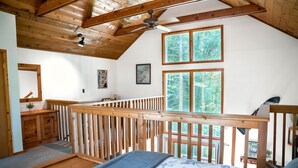 Een slaapkamer met een houten plafond, een bed met een bloemenprintdeken, een houten leuning en uitzicht op bomen door grote ramen.