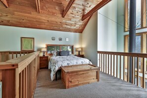 A bedroom with a wooden ceiling, a bed with a floral bedspread, two bedside tables with lamps, a framed picture on the wall, and a window with a view of trees. - Bob's Sled Lodge – 2BR Killington Retreat Near Slopes & Trails (Killington)