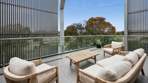 Een balkon met rieten meubilair, een glazen balustrade en uitzicht op bomen en gebouwen.