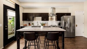 Eine moderne Küche mit dunklen Schränken, einem Edelstahlkühlschrank, einer zentralen Kochinsel mit Barhocker und Blick ins Freie durch eine Glastür.