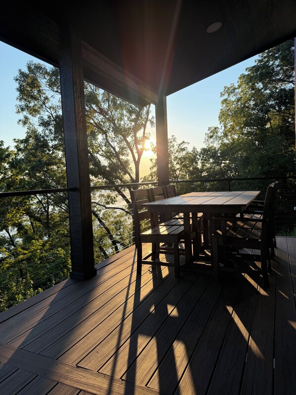 An outdoor wooden deck with a table and chairs, surrounded by trees and a clear sky.