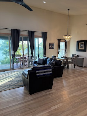 A spacious living room with a black sofa, a dining area with a table and chairs, and a large window with curtains.