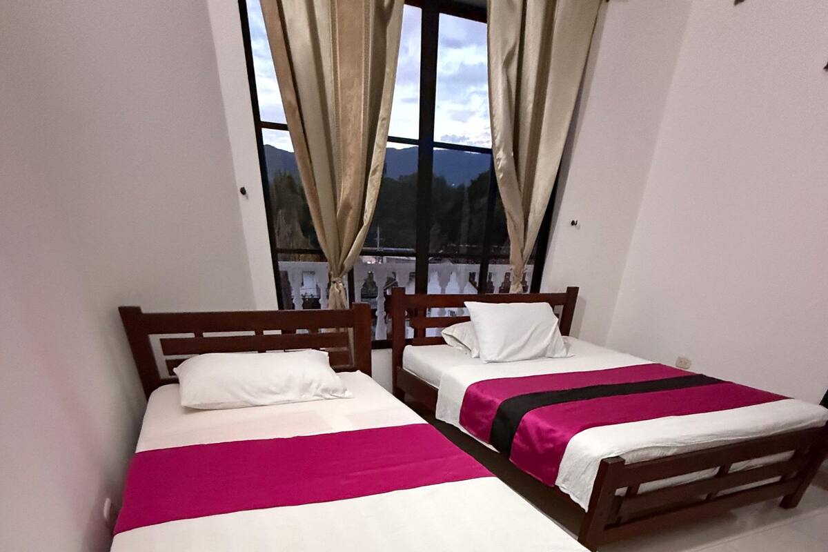 A room with two wooden beds, white bedding, and a view through a window with curtains.