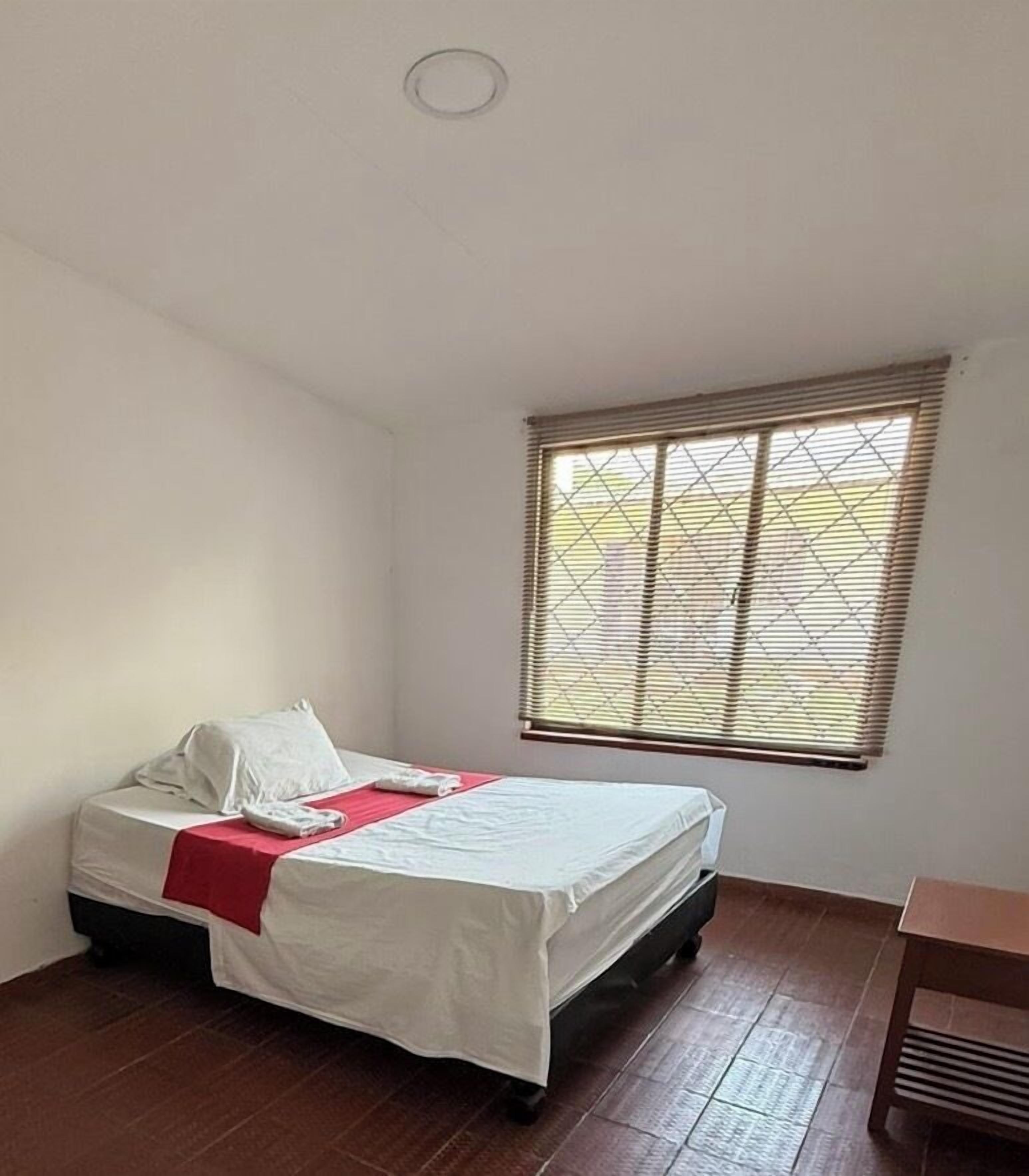 A single bed with a white mattress and red blanket, a window with blinds, and a wooden floor.