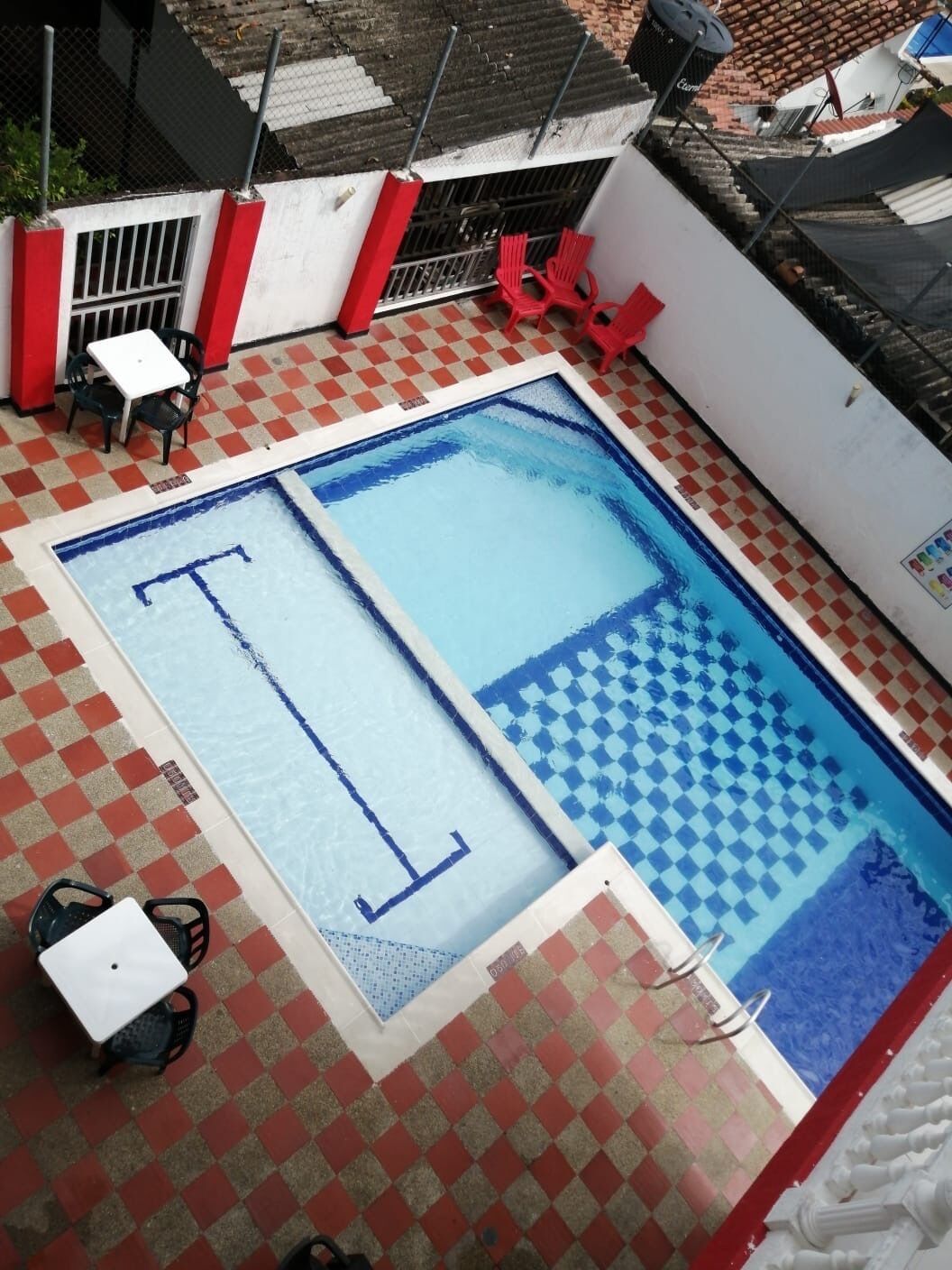 A rectangular pool with a checkered pattern on one side and a blue border. There are red chairs and a white table around the pool.