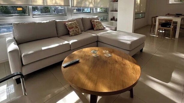 A modern living room with a beige sectional sofa, a wooden coffee table, and large windows with blinds.