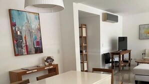A dining area with a white table and wooden chairs, a painting on the wall, and a TV in the background.