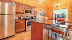 A wooden cabin interior with a kitchen featuring a stainless steel refrigerator, microwave, stove, and oven. There is a bar area with stools and a sign advertising vacation rentals. - Dancing Bear Hideaway - Hot Tub, Resort Pool & Wooded Mountain Retreat (Gatlinburg)