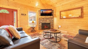 A cozy cabin interior with a fireplace, leather sofas, a TV mounted above it, and a large window with blinds. - Dancing Bear Hideaway - Hot Tub, Resort Pool & Wooded Mountain Retreat (Gatlinburg)