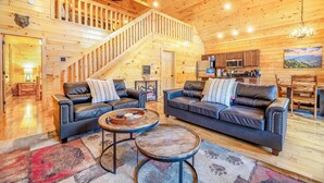 A log cabin interior with two leather sofas, a wooden dining table, a kitchen area, and a staircase. - Dancing Bear Hideaway - Hot Tub, Resort Pool & Wooded Mountain Retreat (Gatlinburg)