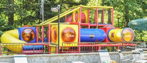 A playground with slides and climbing structures, surrounded by lounge chairs and a stone wall.