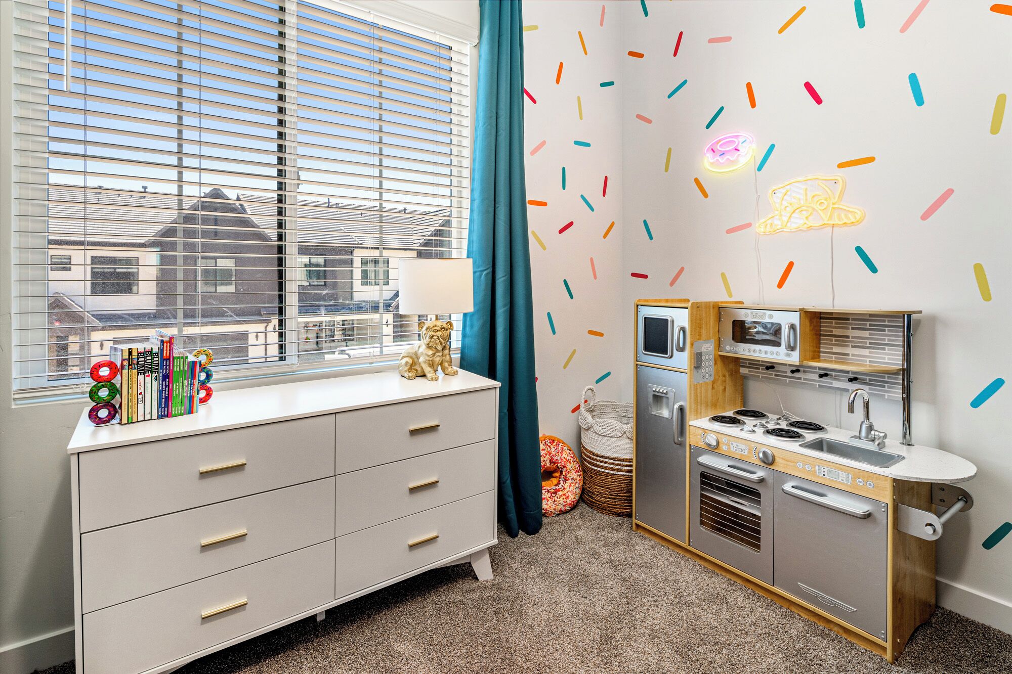 A compact kitchenette with a microwave, stove, and sink, a white dresser with drawers, a window with blinds, and a wall with colorful sprinkles.