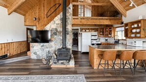 A spacious kitchen with a stone fireplace, wooden cabinets, and a large island with bar stools. - Spectacular views from cabin in Cle Elum. Hiking, fishing, ATVs, and breweries! (Cle Elum)