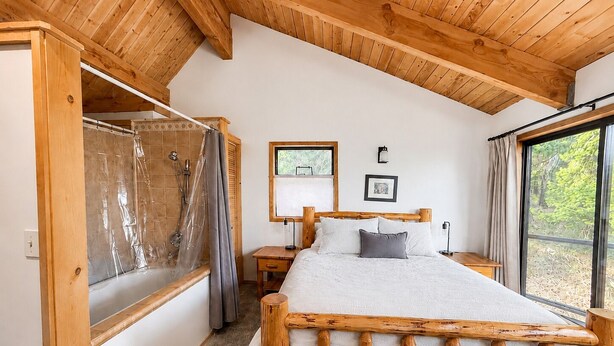 A wooden-framed bed in a room with a shower, a bedside table, and a window with a view of greenery. - Spectacular views from cabin in Cle Elum. Hiking, fishing, ATVs, and breweries! (Cle Elum)