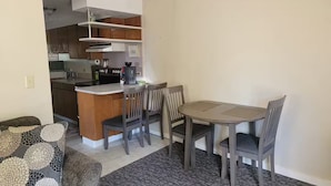 Een knus woongedeelte met een kitchenette, een eethoek met een tafel en vier stoelen, en een grijs gestreept bankje.
