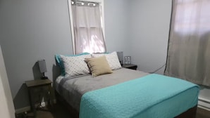 Een slaapkamer met een bed, nachtkastje, lamp, raam met gordijnen en een bord aan de muur.