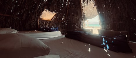 A thatched-roof room with a bed, pillows, and a view of the beach.