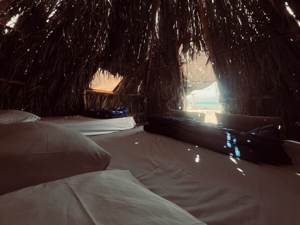 A thatched-roof room with a bed, pillows, and a view of the beach.