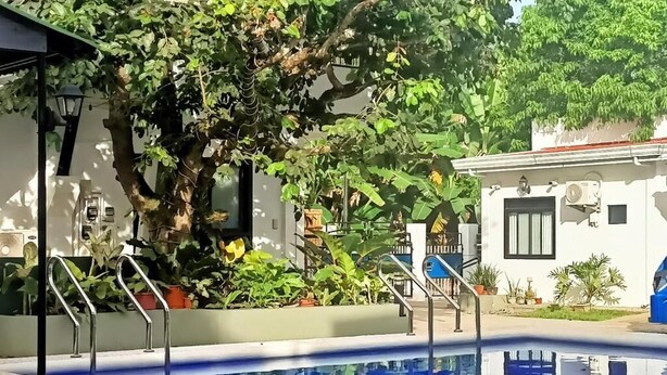 Una piscina con barandas metálicas, rodeada de exuberante follaje y un edificio blanco con techo rojo.