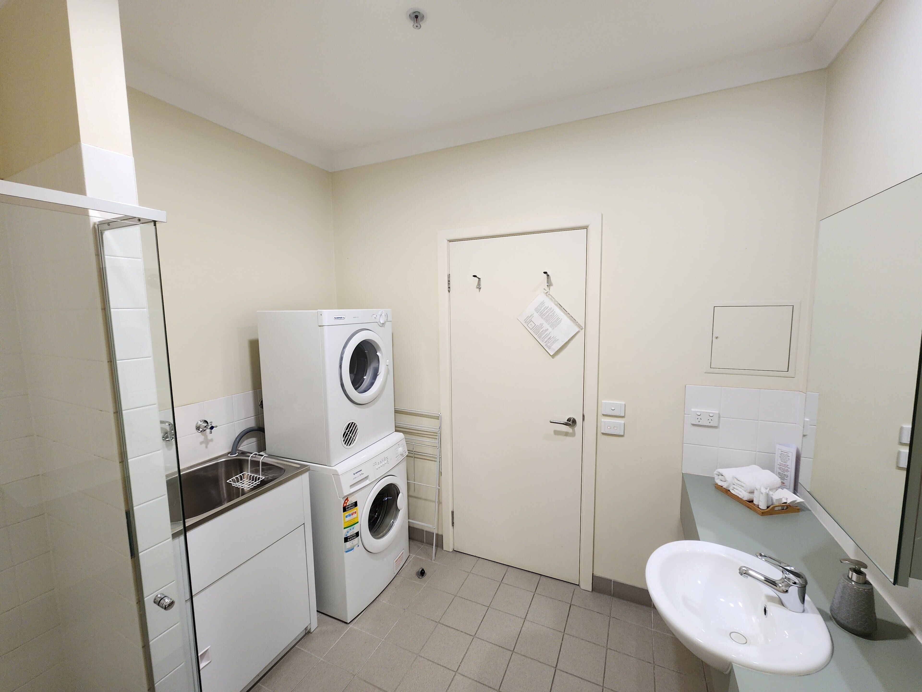 A compact hotel bathroom with a washing machine, a sink, and a mirror.