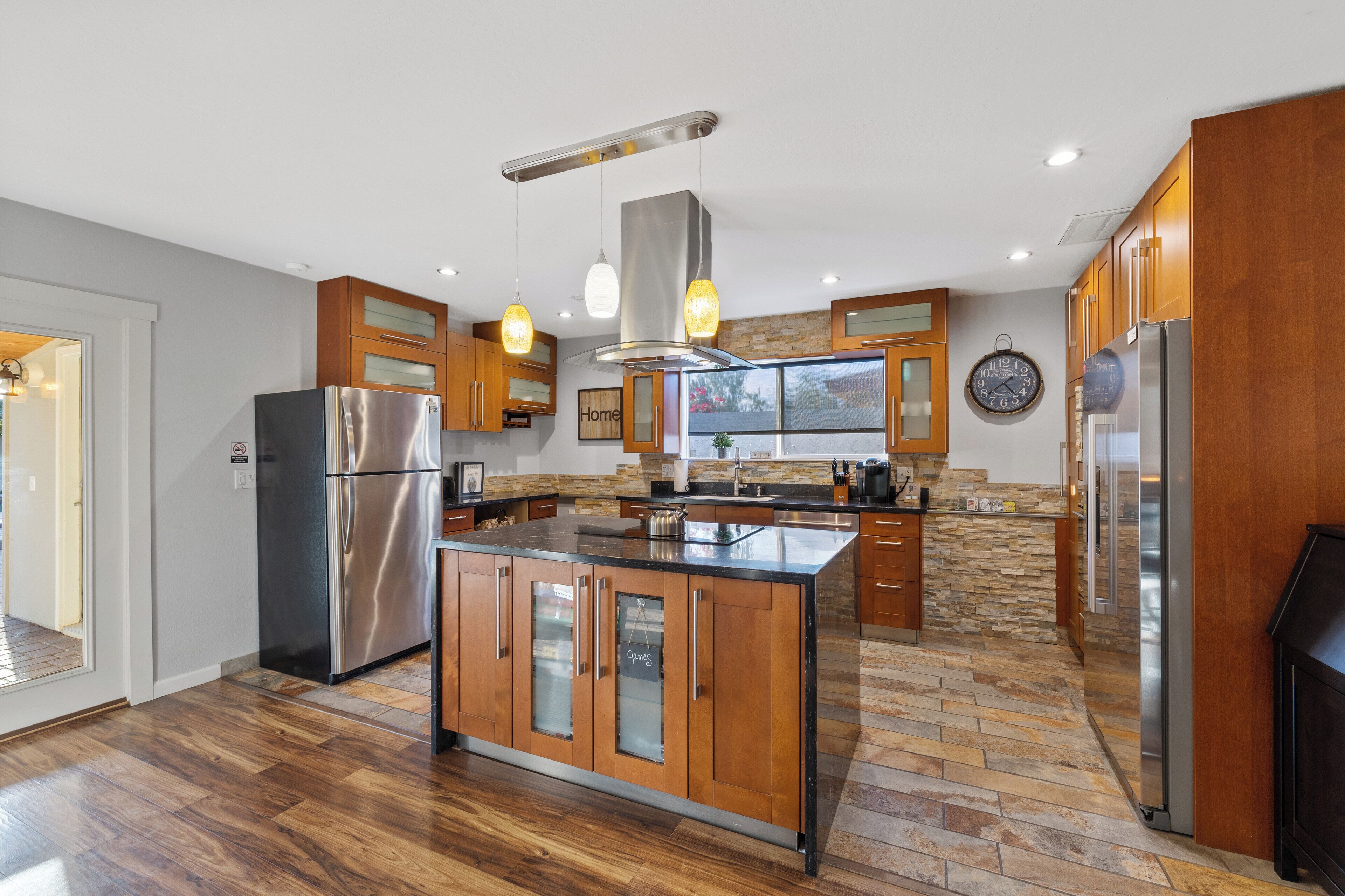 A modern kitchen with a central island, stainless steel appliances, wooden cabinets, and a stone backsplash.