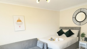 Une chambre avec un canapé gris, un lit avec des oreillers, une table de chevet, un miroir fixé au mur et un luminaire au plafond.