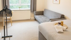 Une chambre d’hôtel avec un lit, un canapé gris, un radiateur, une fenêtre avec des rideaux, une plante et un tableau encadré au mur.