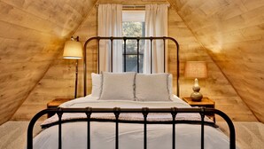 Un dormitorio acogedor en el ático con estructura de cama metálica, una ventana con cortinas y una lámpara en una mesita de noche.