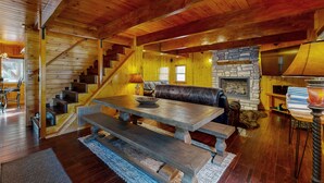 Interior de una cabaña de madera con chimenea de piedra, escalera de madera y comedor con bancos.