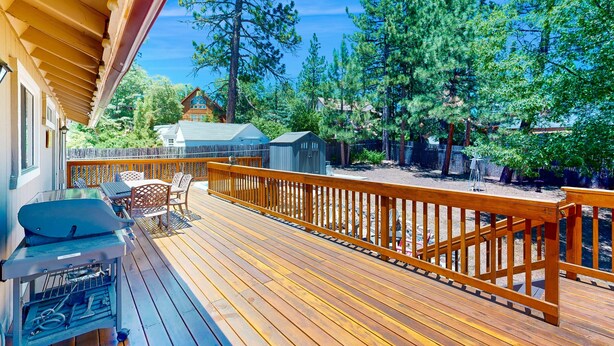 Una terraza de madera con una parrilla, mobiliario exterior y vistas a árboles y edificios circundantes.