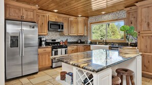A modern kitchen with wooden cabinets, stainless steel appliances, a granite countertop, and a large window offering a view of trees.