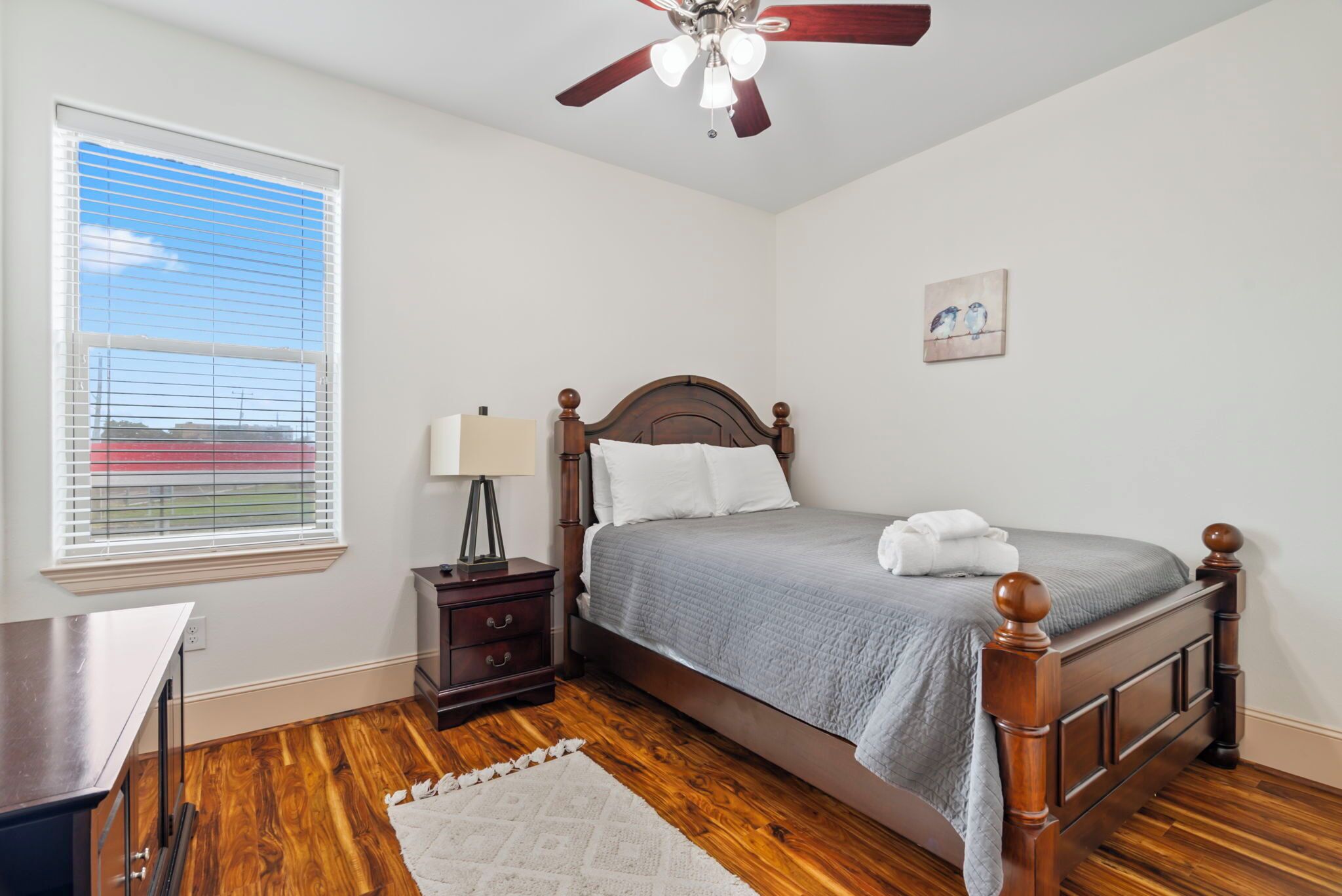 A bedroom with a wooden bed, a nightstand, a desk, a ceiling fan, and a window with blinds.