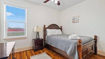 A bedroom with a wooden bed, a nightstand, a desk, a ceiling fan, and a window with blinds.
